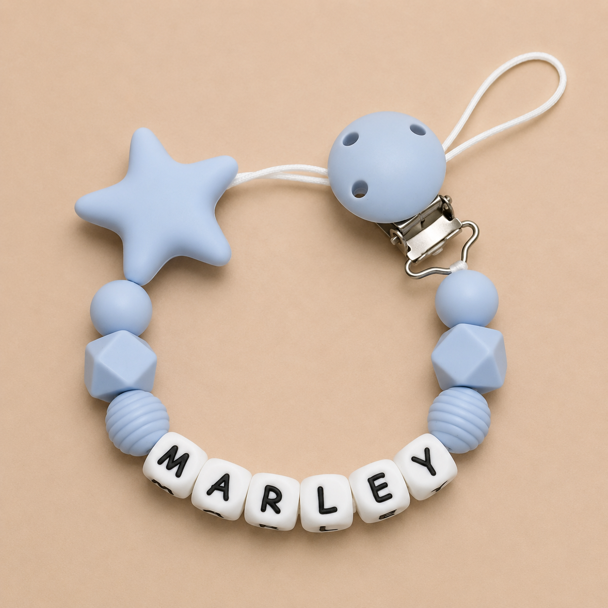 Blue baby dummy clip with star and geometric shapes, featuring the name 'MARLEY' on a beige background.