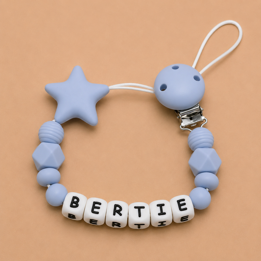 Blue silicone dummy clip with star-shaped bead and 'BERTIE' letter beads on a beige background