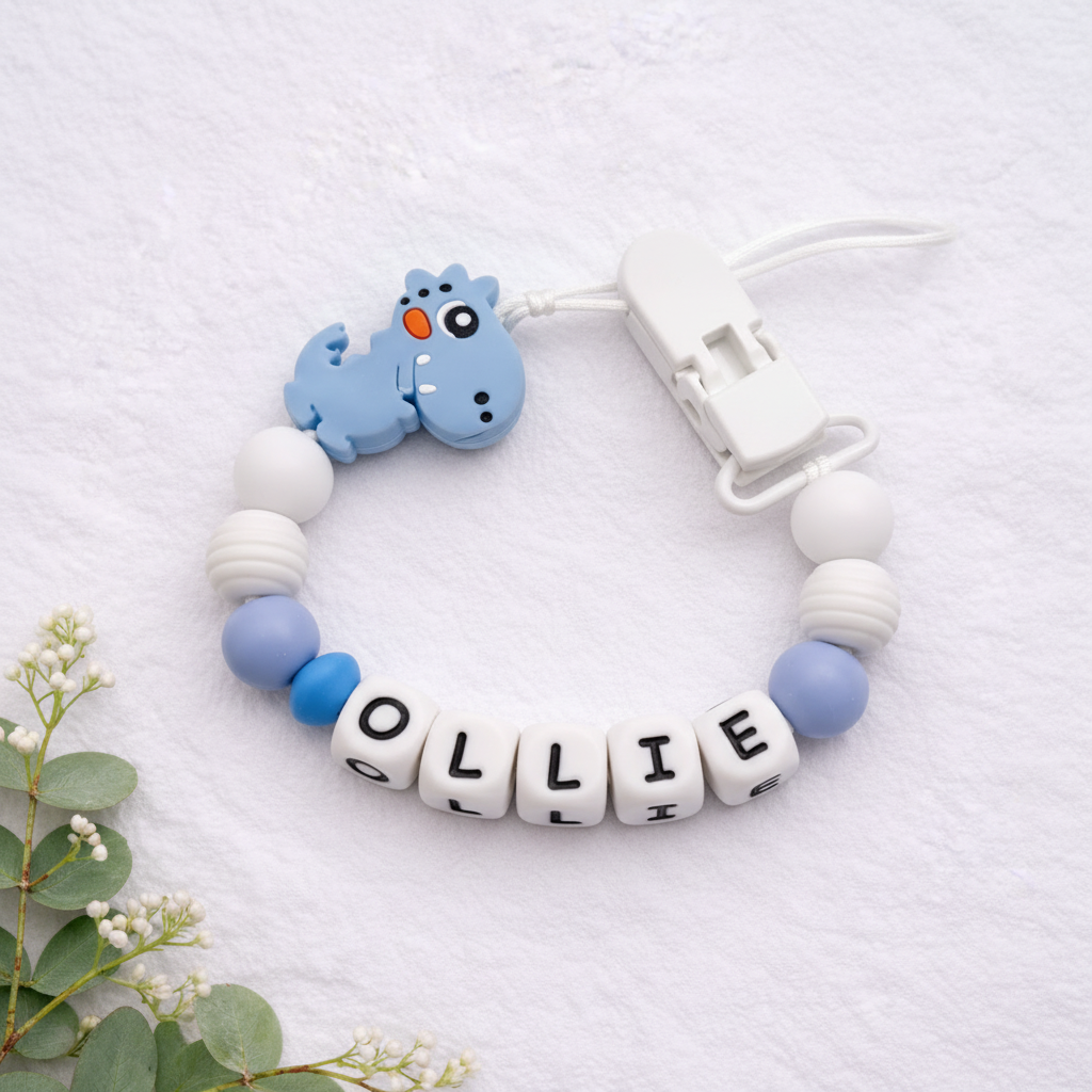 Personalised blue and white dinosaur dummy clip close-up on a soft white luxury textured background.