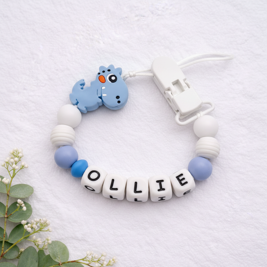 Personalised blue and white dinosaur dummy clip close-up on a soft white luxury textured background.