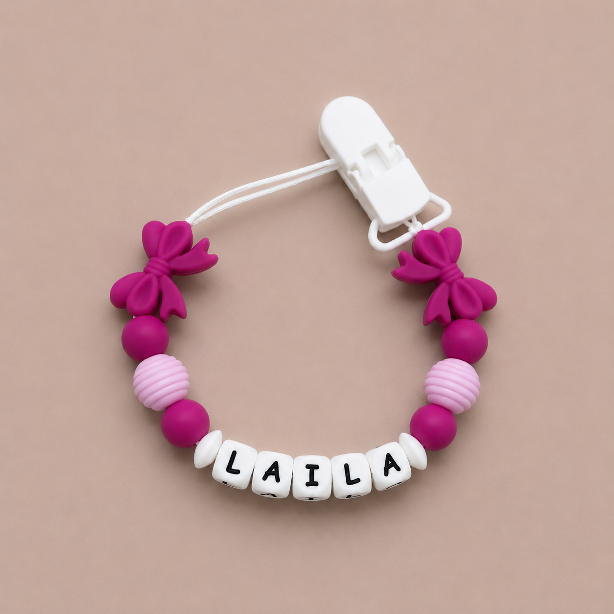 Pink and white pacifier clip with 'LAILA' on a beige background