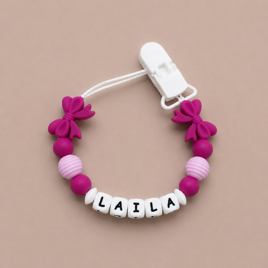 Pink and white pacifier clip with 'LAILA' on a beige background