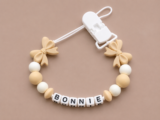 Baby dummy clip with bow-shaped ends and 'BONNIE' letter beads on a beige background