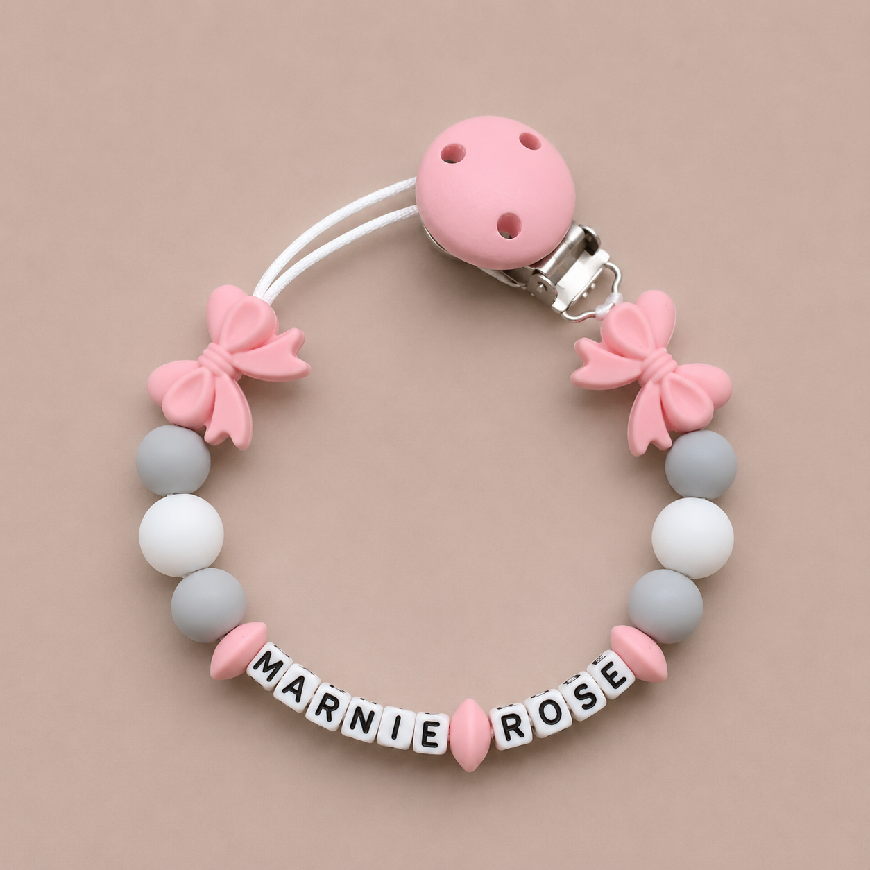 Baby dummy clip with pink and gray beads and 'MARNIE ROSE' letters on a beige background