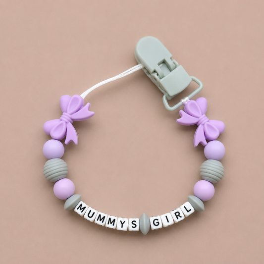 Baby dummy clip with purple and gray beads and 'MUMMY'S GIRL' clasp on a beige background