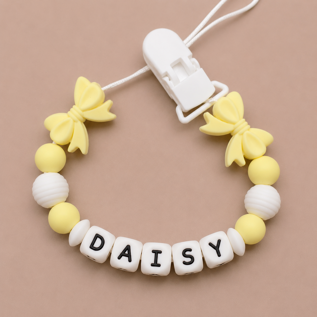 Baby dummy clip with yellow butterfly charms and 'DAISY' letter beads on a beige background
