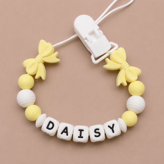 Baby dummy clip with yellow butterfly charms and 'DAISY' letter beads on a beige background