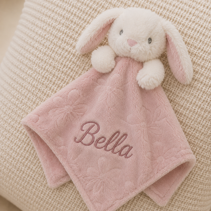 A pink plush baby security blanket with a white bunny head and long floppy ears, embroidered with the name 'Bella' in pink on the front, laid on a textured beige fabric surface.
