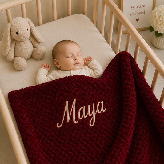 A cosy newborn tucked into a wooden crib under a personalised burgundy bubble blanket embroidered with "Maya" in Gold. A heartwarming keepsake blanket and special gift from Nana.