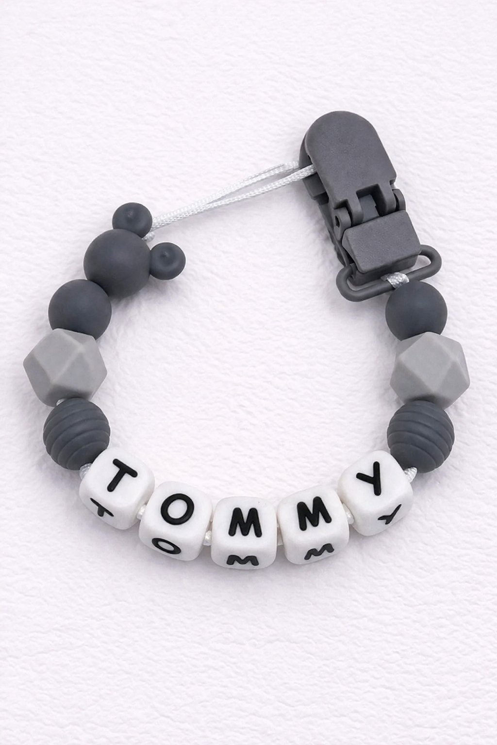 Gray silicone pacifier clip with letter beads spelling 'Tommy' on a white background