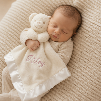 Newborn baby sleeping with a white teddy bear and blanket with 'Riley' embroidered on it, on a beige textured surface.