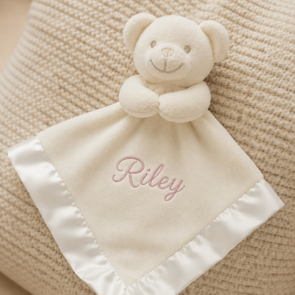 White teddy bear security blanket with 'Riley' embroidered on a textured beige surface