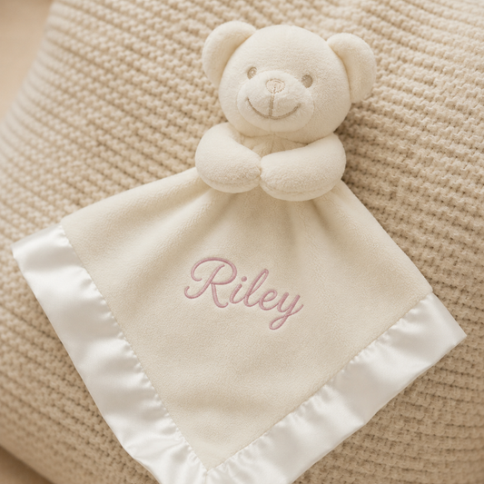 White teddy bear security blanket with 'Riley' embroidered on a textured beige surface