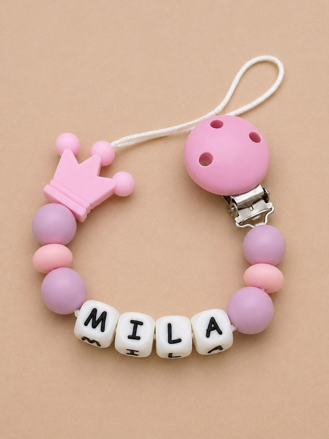 Pink and purple pacifier clip with 'MILA' on a beige background