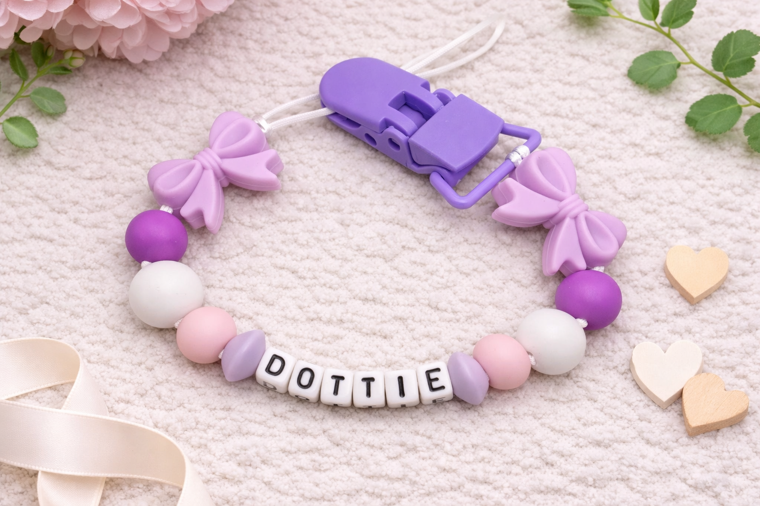 Flat lay of a personalised pacifier clip named “DOTTIE” with lilac bows, purple and pastel beads, and a purple clip displayed on a soft cream textured background with decorative flowers and wooden hearts.