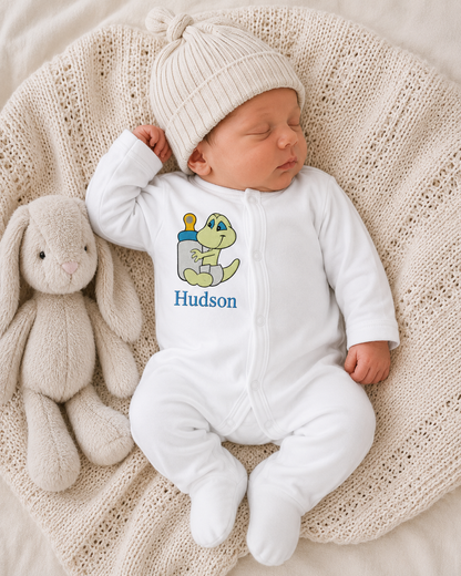 Newborn baby in a white onesie with a frog design and 'Hudson' text, wearing a matching hat, lying on a textured blanket with a plush toy.