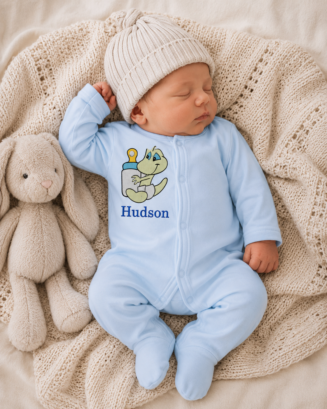 Newborn baby in a blue onesie with a cartoon design and name, lying on a beige textured blanket with a plush toy.