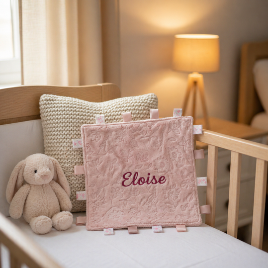Pink baby blanket with 'Eloise' embroidered, placed on a crib with a soft toy and pillow.