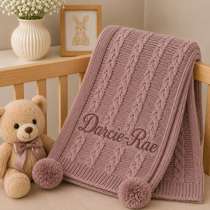 A dusty pink cable-knit baby blanket with two pom-poms and the name 'Darcie-Rae' embroidered on it, styled on a wooden crib next to a teddy bear and a small bunny picture frame.