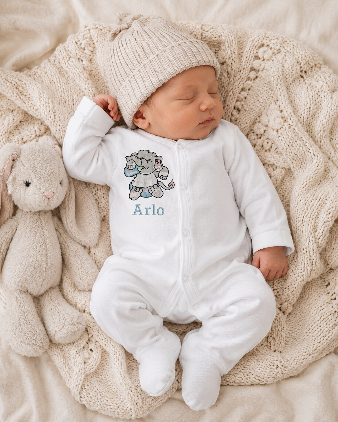 Newborn baby in a white onesie with a cartoon elephant and 'Arlo' text, wearing a matching hat, surrounded by soft textures.