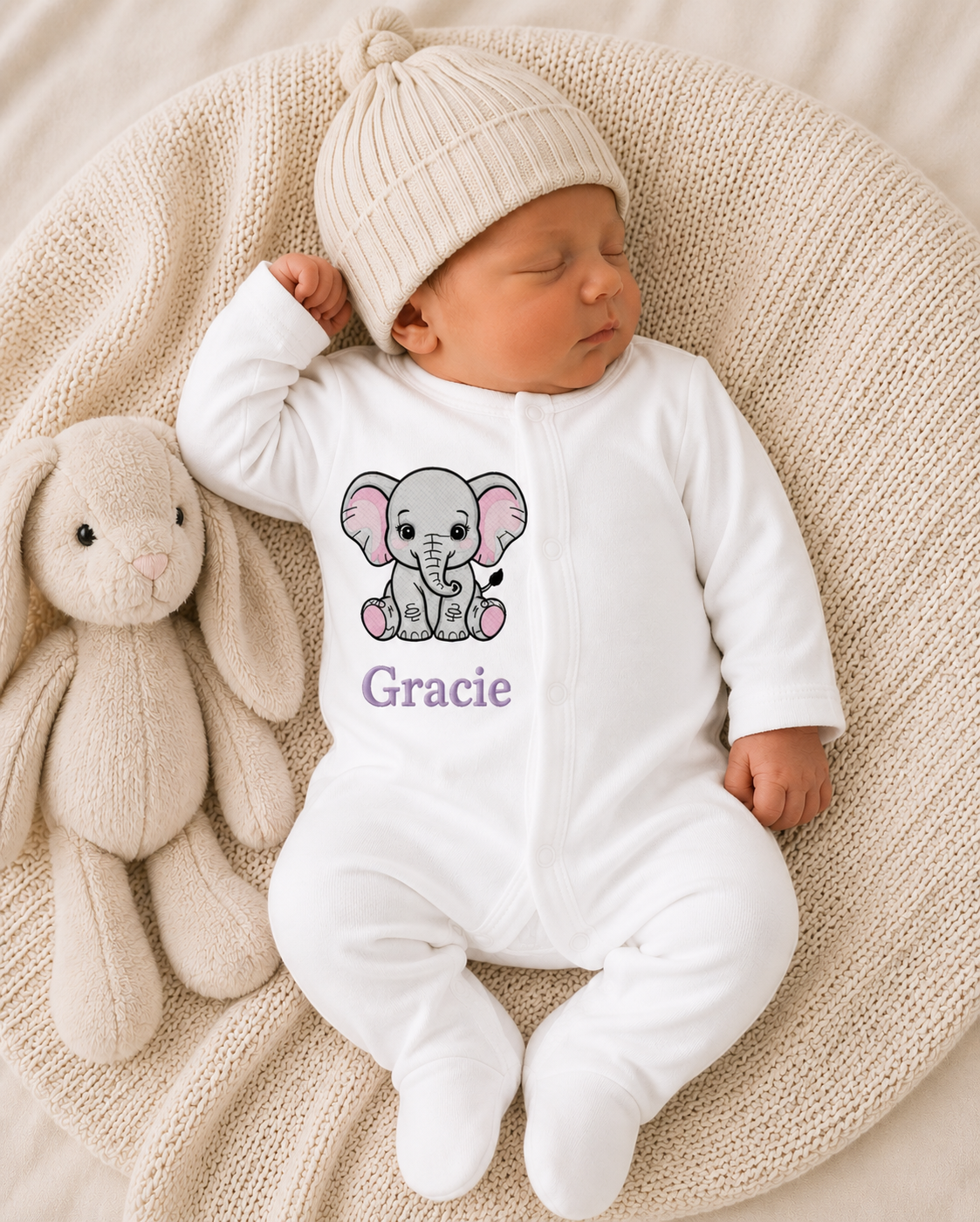 Newborn baby in a white onesie with an elephant design and 'Gracie' text, next to a plush toy on a textured surface.