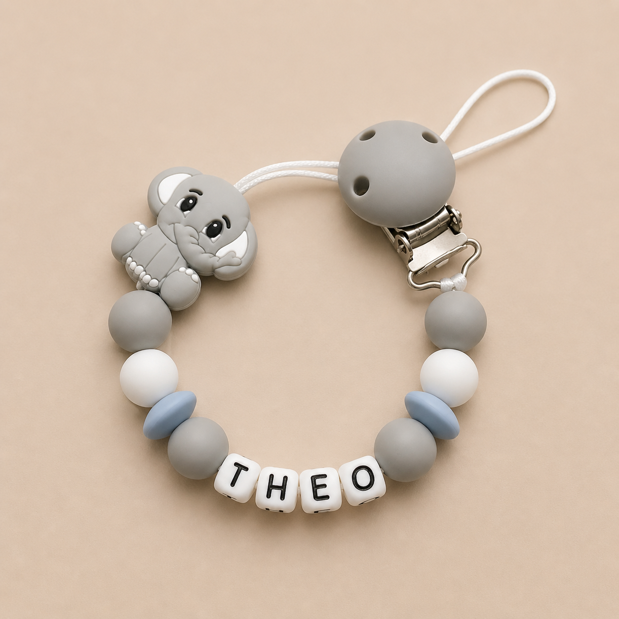 Silicone dummy clip with elephant charm and 'THEO' beads on a beige background
