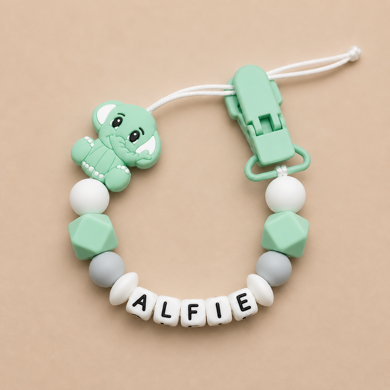dummy clip with dinosaur charm and 'ALFIE' beads on a beige background