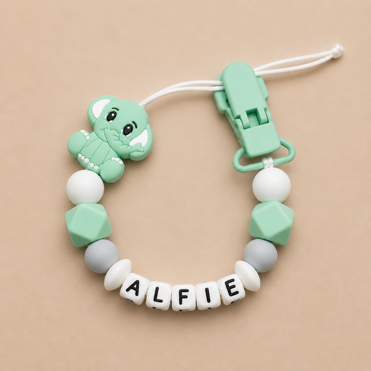 dummy clip with dinosaur charm and 'ALFIE' beads on a beige background