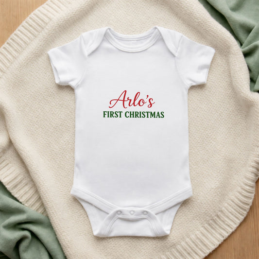 White baby onesie with 'Arlo's First Christmas' text on a textured surface