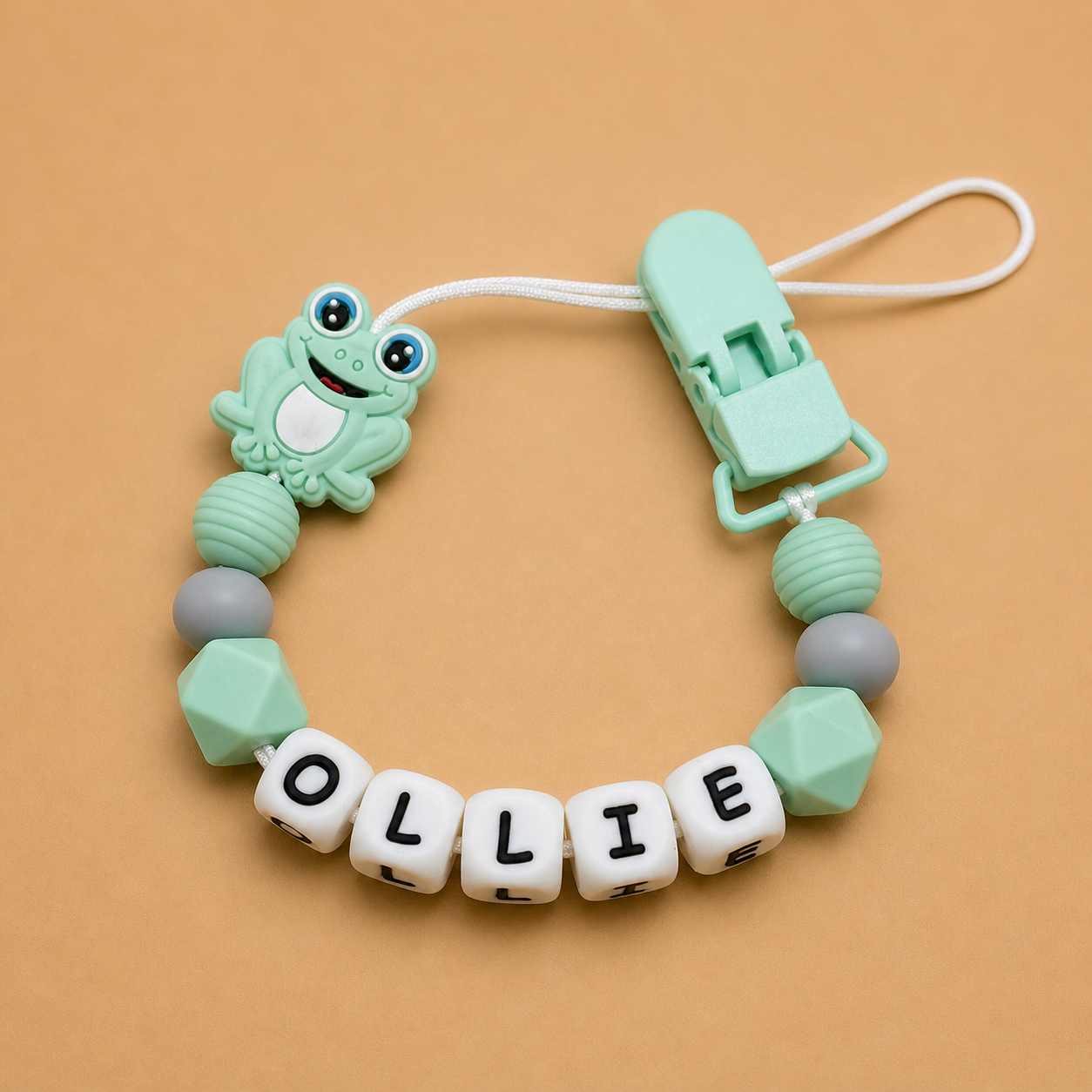 dummy clip with frog design and 'OLLIE' beads on a beige background