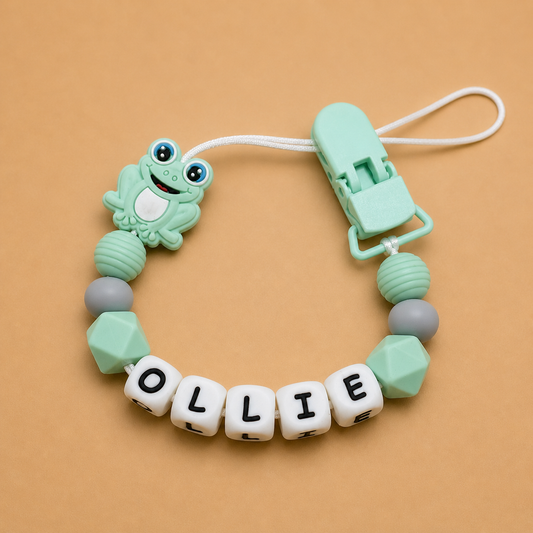 dummy clip with frog design and 'OLLIE' beads on a beige background