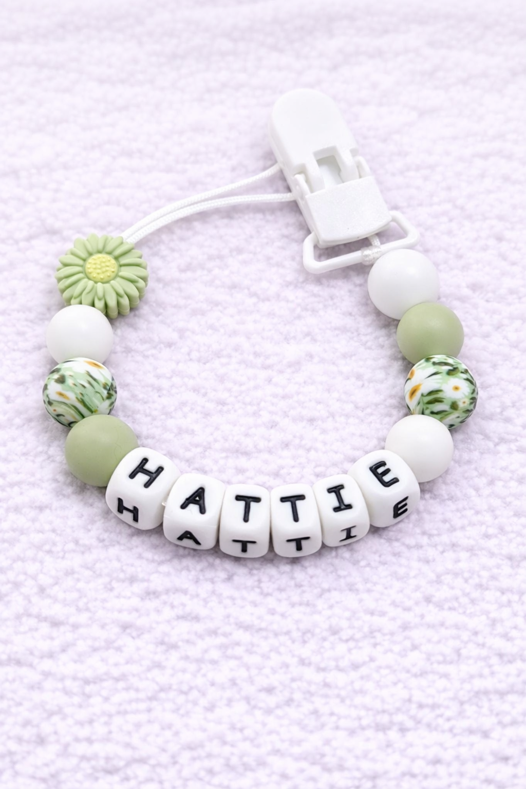 Daisy pacifier clip with 'HATTIE' on a light purple textured background