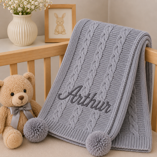 A light gray cable-knit baby blanket draped over a wooden crib, personalized with the name 'Arthur' in dark gray script and finished with two pom-poms. A beige teddy bear and a white vase with flowers are placed beside it in a nursery setting.