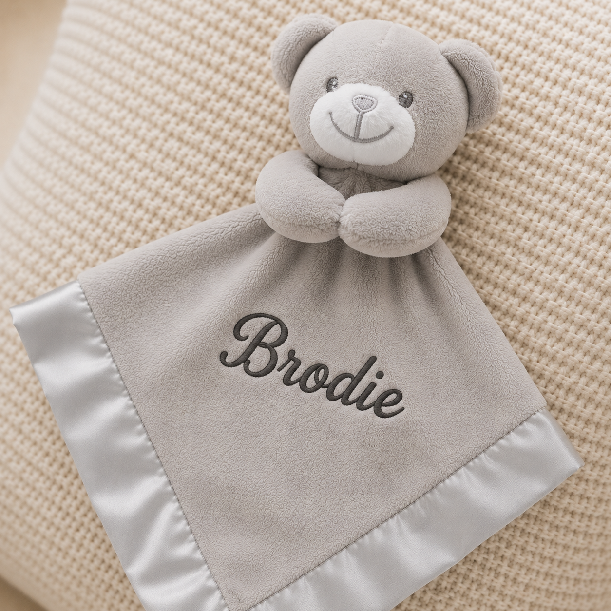 Gray teddy bear security blanket with 'Brodie' embroidered on it, placed on a textured beige surface.