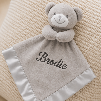 Gray teddy bear security blanket with 'Brodie' embroidered on it, placed on a textured beige surface.