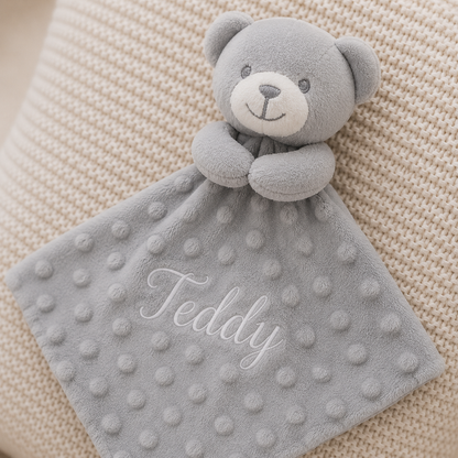 Gray teddy bear security blanket with 'Teddy' embroidery on a beige textured background