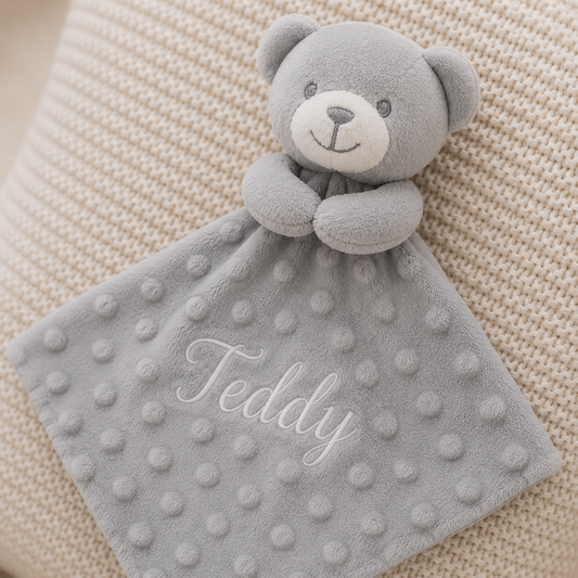 Gray teddy bear security blanket with 'Teddy' embroidery on a beige textured background