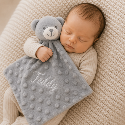 Newborn baby holding a gray teddy bear and blanket with 'Teddy' on a beige textured surface