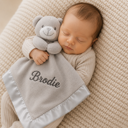 Newborn baby holding a gray teddy bear and blanket with 'Brodie' embroidered, lying on a textured beige surface.
