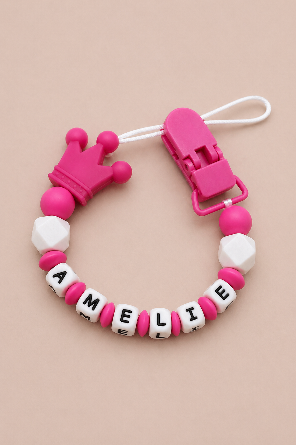 Pink and white pacifier clip with letter beads spelling 'AMELIE' on a beige background