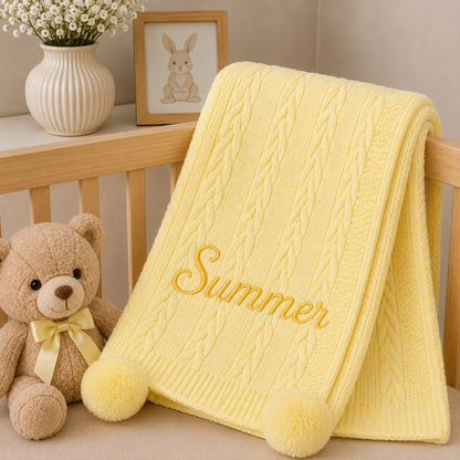 A pale yellow cable-knit baby blanket with 'Summer' embroidered in yellow script, draped over a wooden crib next to a teddy bear and a small bunny picture frame.