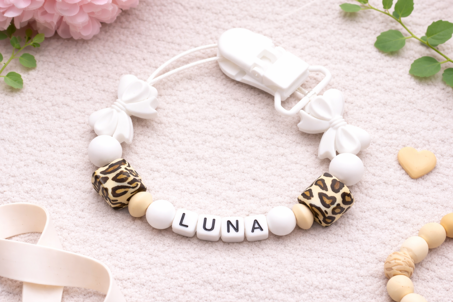 Flat lay photograph of a personalised baby dummy clip spelling “LUNA,” featuring white silicone beads, leopard print accent beads, and white bow details. The clip is arranged on a soft cream textured background with pink flowers, green leaves, and small wooden heart props for a styled product mock-up.