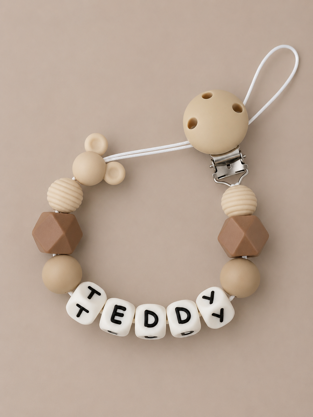 A personalized baby pacifier clip featuring beige and brown silicone beads, a white clip, and white letter beads spelling 'TEDDY' on a warm beige background.