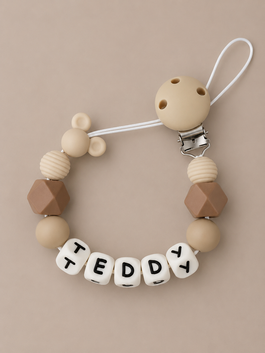 A personalized baby pacifier clip featuring beige and brown silicone beads, a white clip, and white letter beads spelling 'TEDDY' on a warm beige background.