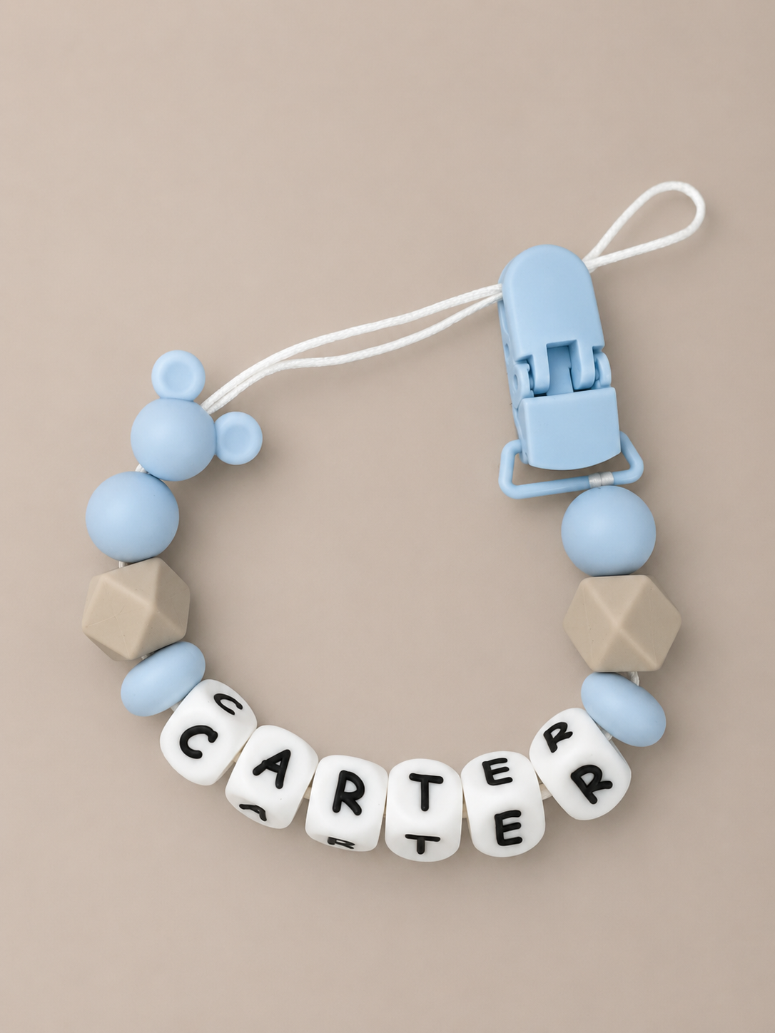A baby teether or clip accessory with light blue silicone beads, beige geometric beads, and white letter beads spelling 'CARTER' on a beige background.