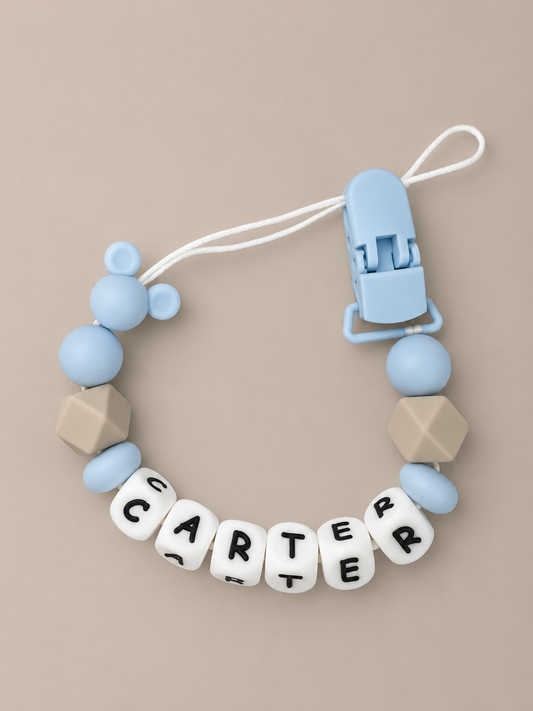 A baby teether or clip accessory with light blue silicone beads, beige geometric beads, and white letter beads spelling 'CARTER' on a beige background.
