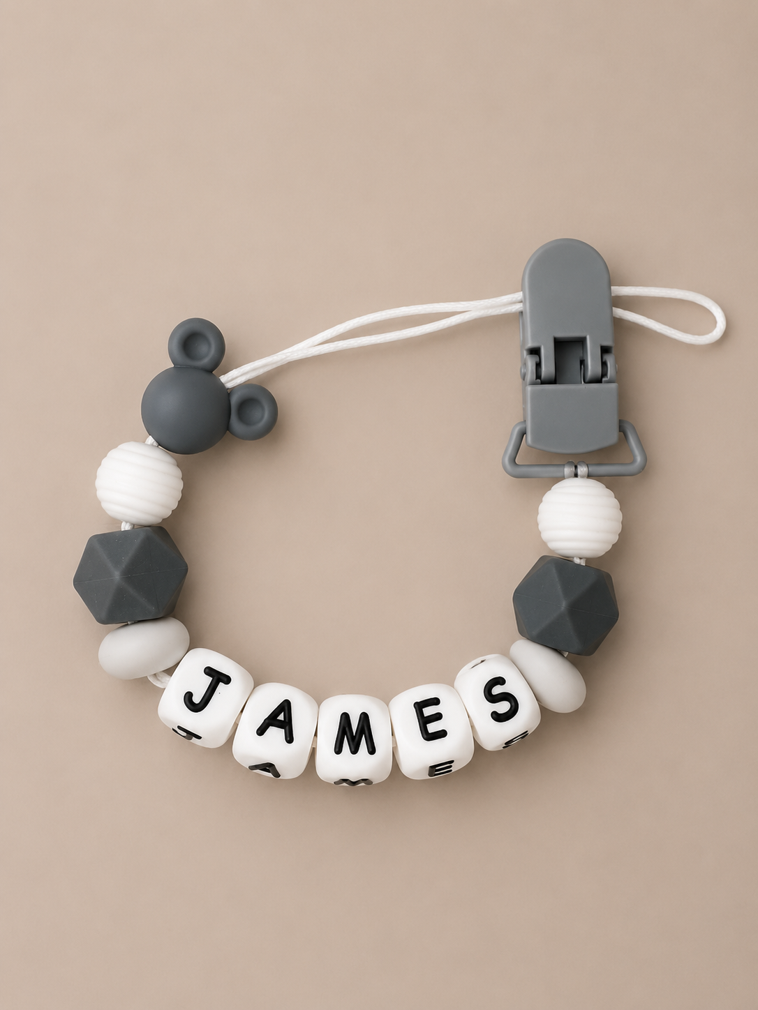 A personalized baby dummy clip featuring white and dark gray silicone beads, including a Mickey Mouse silhouette, faceted geometric beads, and lettered beads spelling 'JAMES'.
