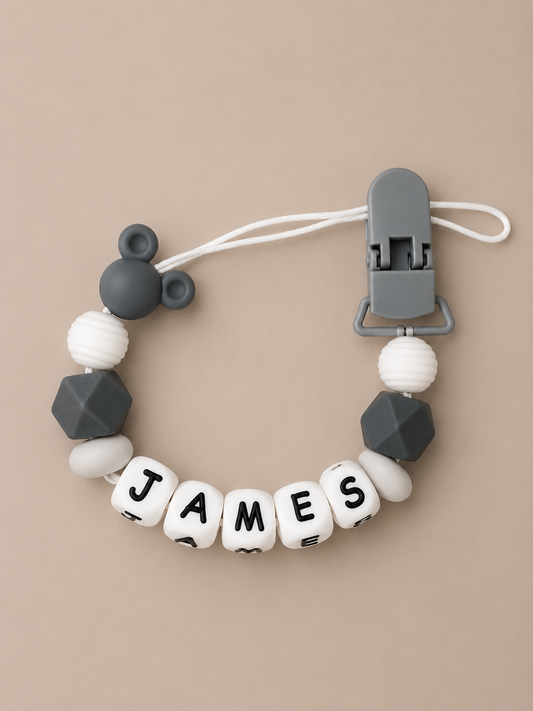 A personalized baby dummy clip featuring white and dark gray silicone beads, including a Mickey Mouse silhouette, faceted geometric beads, and lettered beads spelling 'JAMES'.