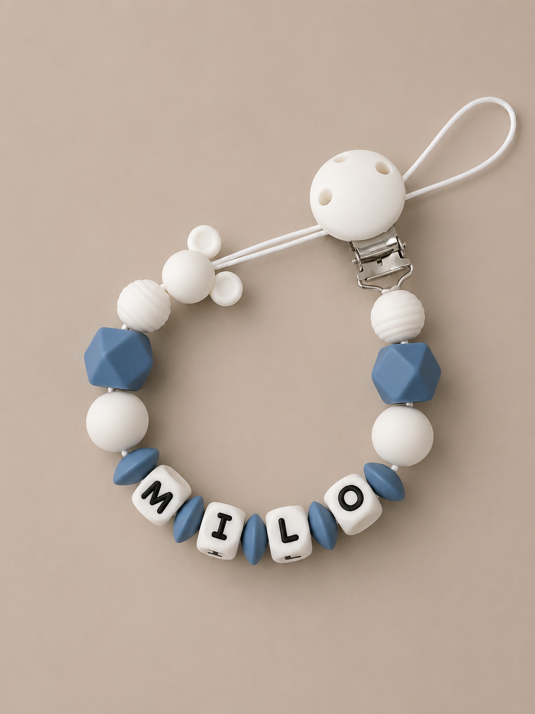 A baby pacifier clip made of white and blue silicone beads, featuring letter beads spelling 'MILo' on a beige background.