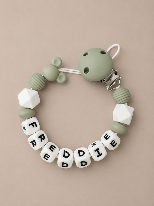 A silicone baby pacifier clip bracelet on a warm beige background. The bracelet features sage green silicone beads, textured green beads, white geometric beads, and white letter beads spelling 'FREDDE' with black letters.